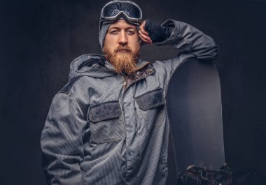 Bir kış şapka ve koruyucu gözlük tam sakallı vahşi kızıl saçlı snowboard snowboard bir stüdyoda ile poz bir snowboard ceket giymiş. Gri bir arka plan üzerinde izole.