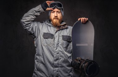 Bir kış şapka ve koruyucu gözlük tam sakallı vahşi kızıl saçlı snowboard snowboard bir stüdyoda ile poz bir snowboard ceket giymiş. Gri bir arka plan üzerinde izole.