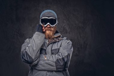 Uzağa arıyorsunuz bir dalgın Kızıl saçlı snowboard kış şapka ve koruyucu gözlük bir stüdyoda poz bir snowboard ceket giymiş tam sakallı portresi. Karanlık dokulu bir arka plan üzerinde izole.