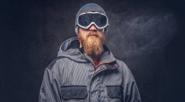 Bir kış şapka ve koruyucu gözlük tam sakallı bir kızıl saçlı snowboard portresi bir stüdyoda poz bir snowboard ceket giymiş. Gri arka plan üzerinde izole.