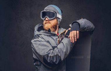 Kızıl saçlı acımasız snowboard uzağa arıyorsunuz bir kış şapka ve koruyucu gözlük bir stüdyoda snowboard ile poz bir snowboard ceket giymiş tam sakallı. Gri bir arka plan üzerinde izole.