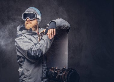 Redhead brutal snowboarder with a full beard in a winter hat and protective glasses dressed in a snowboarding coat posing with snowboard at a studio, looking away. Isolated on a gray background. clipart