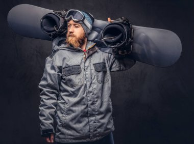 Kızıl saçlı sakallı uzağa arıyorsunuz snowboard kış şapka ve koruyucu gözlük bir stüdyoda snowboard ile poz bir snowboard ceket giymiş. Gri bir arka plan üzerinde izole.