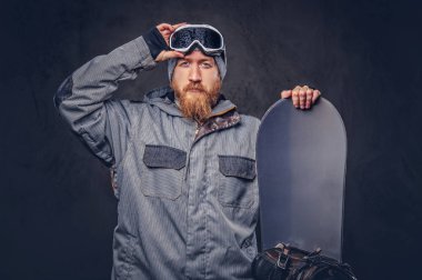 Bir kış şapka ve koruyucu gözlük tam sakallı vahşi kızıl saçlı snowboard snowboard bir stüdyoda ile poz bir snowboard ceket giymiş. Gri bir arka plan üzerinde izole.