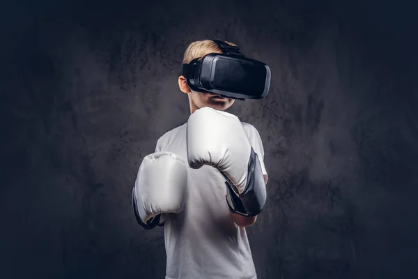Schoolboy boxer with blonde hair dressed in a white t-shirt wearing visual reality glasses and boxing gloves, workout in a studio. Isolated on a dark textured background.