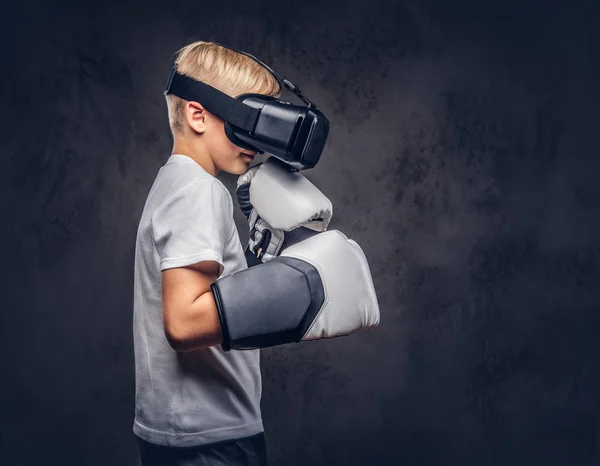 Young boy boxer with blonde hair dressed in a white t-shirt wearing visual reality glasses and boxing gloves, workout in a studio. Isolated on a dark textured background.