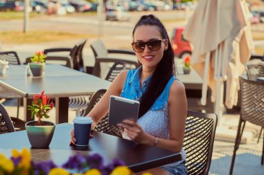 Bir tablet bir terasta açık havada kafe, kameraya bakarak otururken yaz günü zevk trendy kıyafetler ve güneş gözlüğü takan bir mutlu güzel blogger kız portresi tutar.