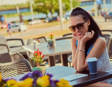 Trendy kıyafetler giymiş mutlu çekici esmer kız portresi yaz günü bir terasta açık havada kafe, kameraya bakarak otururken keyif alıyor.