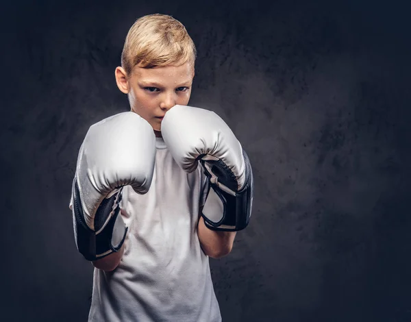 A handsome little boy boxer with blonde hair dressed in a white t-shirt in gloves ready to fight. Isolated on the dark textured background.