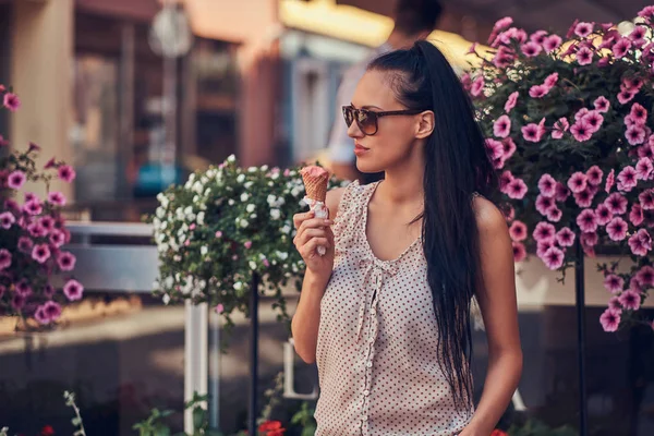Sensual brunette girl in sunglasses wearing trendy clothes is enjoying summer day holds a strawberry ice cream while stands near terrace decorated with flowers.