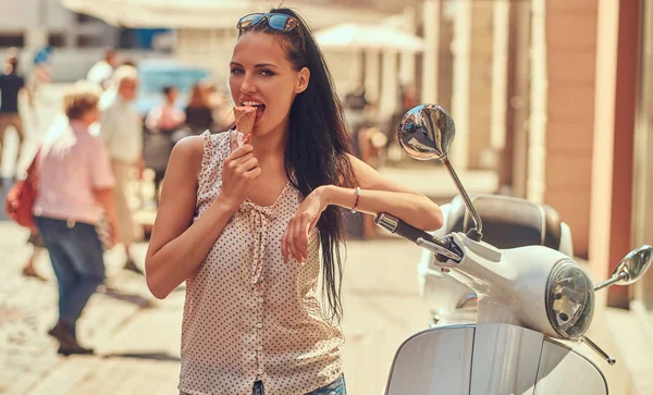 Sensual brunette girl in sunglasses wearing trendy clothes is enjoying a summer day, eating strawberry ice cream while posing on a street near a white classical Italian scooter.