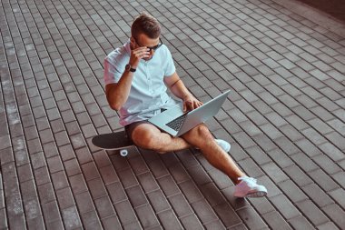 Beyaz bir gömlek ve şort köprü altında bir kaykay üzerinde otururken bir dizüstü bilgisayarda çalışma portre yakışıklı bir moda Freelancer güneş gözlüğü giymiş.