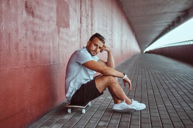 Dalgın moda yakışıklı olan beyaz bir gömlek ve şort köprünün altından bir kaykay üzerinde oturan bir kameraya bakarak giymiş.