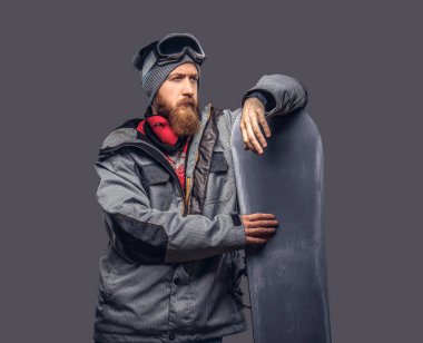 Kızıl saçlı acımasız snowboard uzağa arıyorsunuz bir kış şapka ve koruyucu gözlük bir stüdyoda snowboard ile poz bir snowboard ceket giymiş tam sakallı. Gri bir arka plan üzerinde izole.