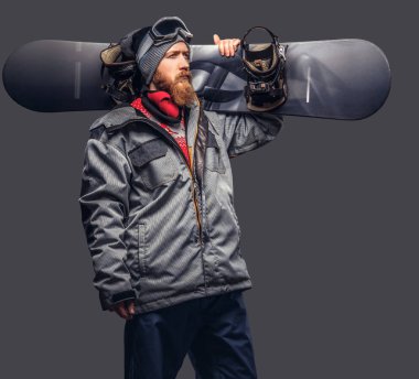 Bir kış şapka ve koruyucu gözlük tam sakallı vahşi kızıl saçlı snowboard snowboard bir stüdyoda ile poz bir snowboard ceket giymiş. Gri bir arka plan üzerinde izole.