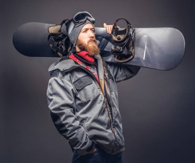 Bir kış şapka ve koruyucu gözlük tam sakallı vahşi kızıl saçlı snowboard snowboard bir stüdyoda ile poz bir snowboard ceket giymiş. Gri bir arka plan üzerinde izole.