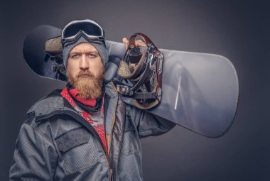 Vahşi kızıl saçlı snowboard kış şapka ve koruyucu gözlük tam sakallı bir stüdyoda bir snowboard ile poz bir snowboard ceket bir kameraya bakarak giymiş. Gri bir arka plan üzerinde izole.