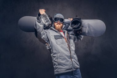 Kızıl saçlı snowboard kış şapka ve koruyucu gözlük bir snowboard ceket giymiş tam sakallı bir stüdyoda onun omzuna bir snowboard ve bir şişe alkol tutar. Gri arka plan üzerinde izole.