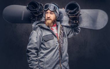 Bir kış şapka ve koruyucu gözlük bir snowboard ceket giymiş tam sakallı vahşi kızıl saçlı snowboard snowboard onun omuz bir stüdyoda tutar. Gri arka plan üzerinde izole.