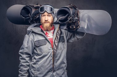 Bir kış şapka ve koruyucu gözlük bir snowboard ceket giymiş tam sakallı vahşi kızıl saçlı snowboard snowboard onun omuz bir stüdyoda tutar. Gri arka plan üzerinde izole.