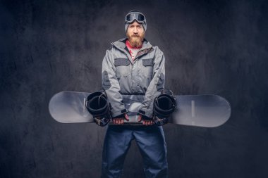 Bir kış şapka ve koruyucu gözlük bir snowboard ceket giymiş tam sakallı vahşi kızıl saçlı snowboard snowboard bir stüdyoda tutar. Karanlık dokulu bir arka plan üzerinde izole.