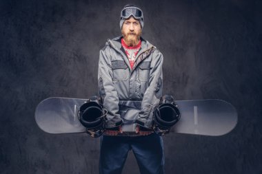 Bir kış şapka ve koruyucu gözlük bir snowboard ceket giymiş Kızıl saçlı sakallı snowboard snowboard bir stüdyoda tutar. Karanlık dokulu bir arka plan üzerinde izole.