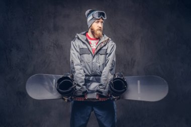 Bir kış şapka ve koruyucu gözlük bir snowboard ceket giymiş Kızıl saçlı sakallı snowboard snowboard bir stüdyoda tutar. Karanlık dokulu bir arka plan üzerinde izole.