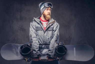 Bir kış şapka ve koruyucu gözlük bir snowboard ceket giymiş tam sakallı vahşi kızıl saçlı snowboard snowboard bir stüdyoda tutar. Karanlık dokulu bir arka plan üzerinde izole.
