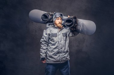 Bir kış şapka ve koruyucu gözlük tam sakallı vahşi kızıl saçlı snowboard snowboard bir stüdyoda ile poz bir snowboard ceket giymiş. Gri bir arka plan üzerinde izole.