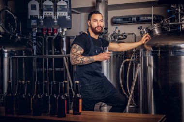 Portrait of a stylish bearded tattooed dark skinned male holds a glass of beer, standing behind the counter in the brewery clipart