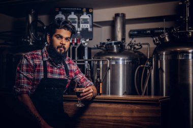 Şık tam Hintli bir yün gömlekli sakallı ve önlük bir bardak bira, bira fabrikası sayacında arkasında oturan tutar.