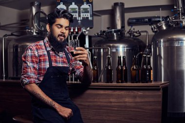 Şık tam Hintli bir yün gömlekli sakallı ve önlük bir bardak bira, bira fabrikası sayacında arkasında oturan tutar.