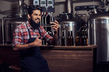Şık tam Hintli bir yün gömlekli sakallı ve önlük bir bardak bira, bira fabrikası sayacında arkasında oturan tutar.