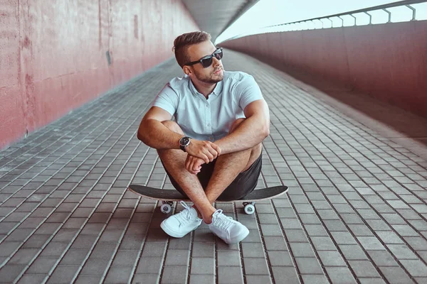 一个英俊的时尚溜冰者的肖像穿着白色衬衫和短裤坐在桥下的滑板上 看着远离 — 图库照片