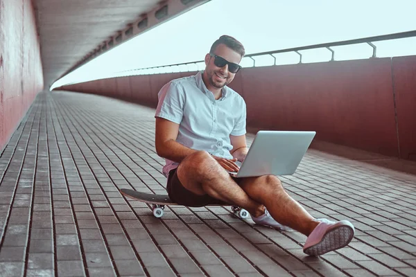 Beyaz bir gömlek ve şort bir kameraya bakarak köprünün altından bir kaykay üzerinde otururken bir dizüstü bilgisayarda çalışan güneş gözlüklü bir mutlu yakışıklı şık serbest meslek portresi giymiş.