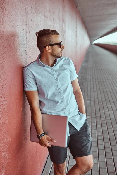 Portre yakışıklı bir moda Freelancer güneş gözlüğü beyaz bir gömlek giymiş ve şort uzağa arıyorsunuz köprünün altından bir duvara yaslanmış bir dizüstü bilgisayar ayakta tutan.