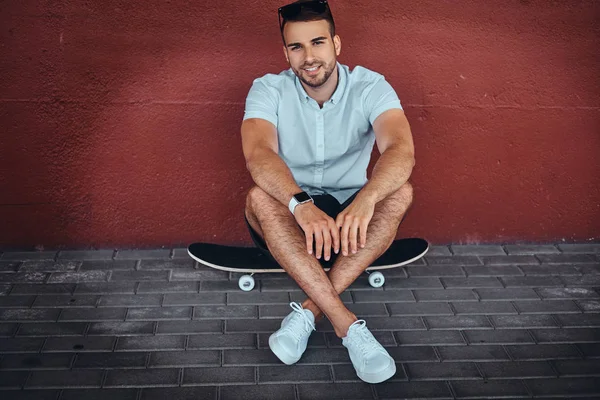 Gülümseyen yakışıklı şık adam beyaz bir gömlek ve şort köprünün altından bir kaykay üzerinde oturan bir kameraya bakarak giymiş.