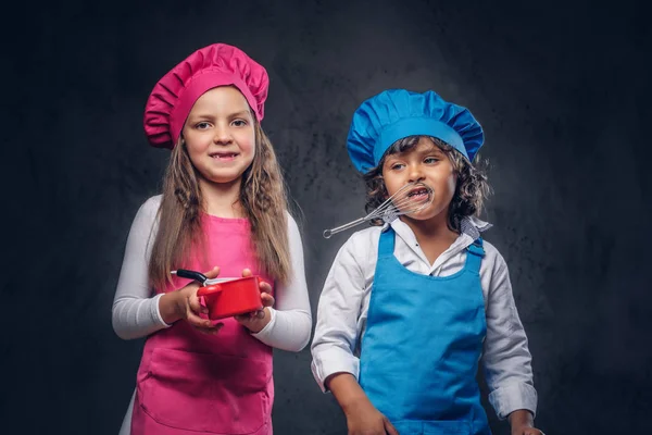 Cute cook couple. Little boy with brown curly hair dressed in a blue cook uniform and beautiful schoolgirl dressed in a pink cook uniform, poses with cookware and fruits at a studio. Isolated on the