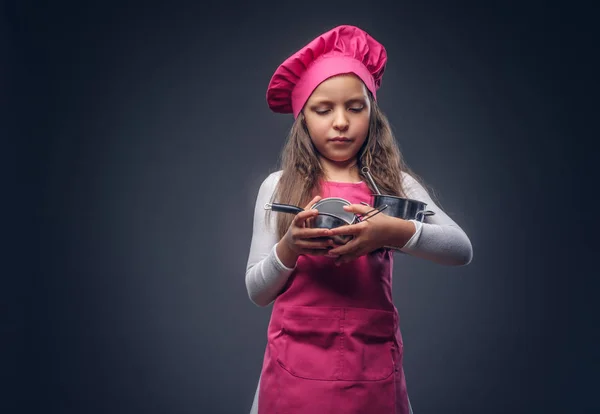 Linda hermosa colegiala vestida con un uniforme de cocinero rosa sostiene utensilios de cocina en un estudio. Aislado sobre el fondo oscuro . —  Fotos de Stock