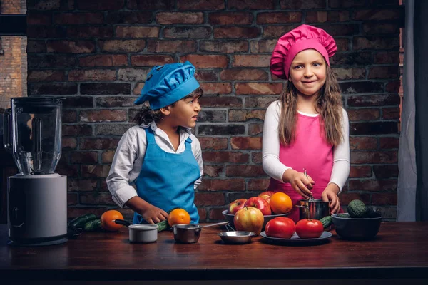 Sevimli aşçı çift. Mavi aşçı üniforma giymiş kahverengi kıvırcık saç ve güzel bir kız bir pembe giymiş olan küçük çocuk yemek Tekdüzen birlikte bir tuğla duvara bir mutfakta yemek.