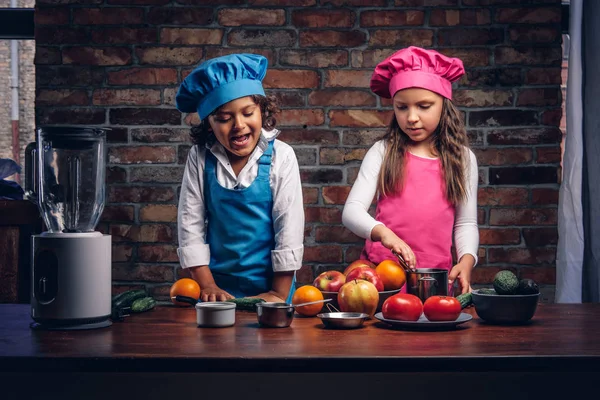 Sevimli aşçı çift. Mavi aşçı üniforma giymiş kahverengi kıvırcık saç ve güzel bir kız bir pembe giymiş olan küçük çocuk yemek Tekdüzen birlikte bir tuğla duvara bir mutfakta yemek.