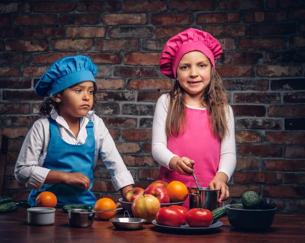 Linda pareja de cocineros. Niño pequeño con el pelo rizado marrón vestido con un uniforme de cocinero azul y una hermosa chica vestida con un uniforme de cocinero rosa cocinando juntos en una cocina contra una pared de ladrillo . —  Fotos de Stock