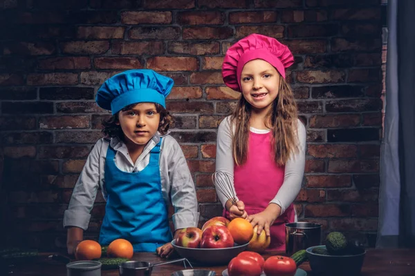Sevimli aşçı çift. Mavi aşçı üniforma giymiş kahverengi kıvırcık saç ve güzel bir kız bir pembe giymiş olan küçük çocuk yemek Tekdüzen birlikte bir tuğla duvara bir mutfakta yemek.