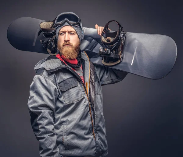 Bir kış şapka ve koruyucu gözlük tam sakallı vahşi kızıl saçlı snowboard snowboard bir stüdyoda ile poz bir snowboard ceket giymiş. Gri bir arka plan üzerinde izole.