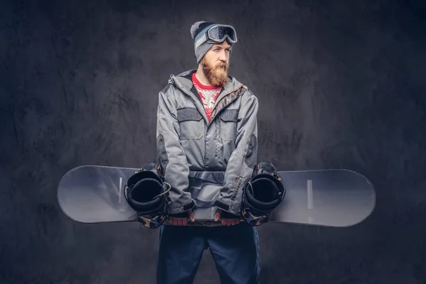 Redhead bearded snowboarder in a winter hat and protective glasses dressed in a snowboarding coat holds a snowboard in a studio. Isolated on a dark textured background.