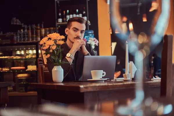 Portrait d'un élégant homme d'affaires avec une barbe élégante et des cheveux vêtus d'un costume noir assis avec la main sur le menton dans un café avec un ordinateur portable ouvert, regardez la caméra . — Photo