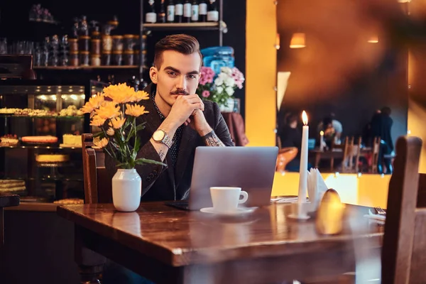 Portret van een peinzende knappe zakenman met stijlvolle baard en haren gekleed in een zwart pak zitten met hand op de kin in een cafe met een opengeklapte laptop, lookong op de camera. — Stockfoto