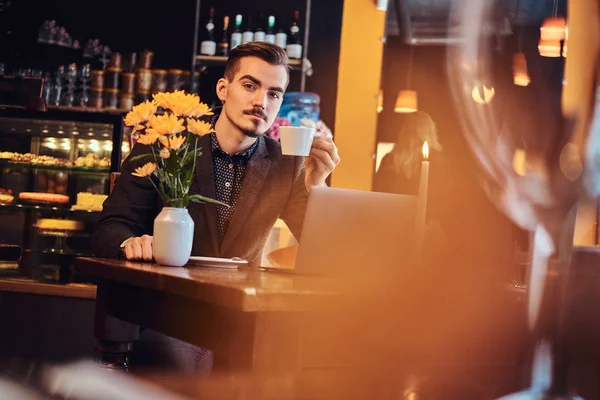 Όμορφος freelancer άνθρωπος με στιλάτα γένια και μαλλιά ντυμένος με ένα μαύρο κοστούμι που κάθεται σε ένα καφέ με ένα ανοιχτό laptop και κατέχει ένα φλιτζάνι καφέ, κοιτάζοντας την κάμερα. — Φωτογραφία Αρχείου