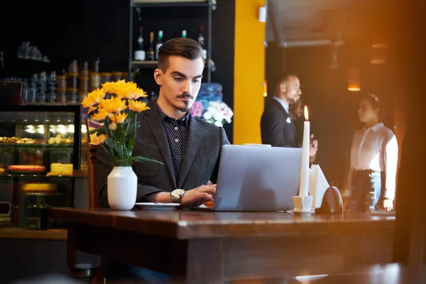 Hombre freelancer guapo con barba elegante y cabello vestido con un traje negro que trabaja en el ordenador portátil mientras está sentado en un café . —  Fotos de Stock