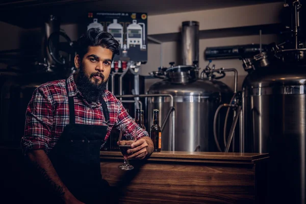 Şık tam Hintli bir yün gömlekli sakallı ve önlük bir bardak bira, bira fabrikası sayacında arkasında oturan tutar.